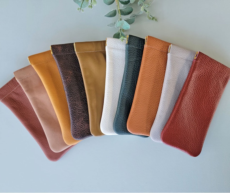 May include: A collection of leather eyeglass cases in various colors, including brown, tan, white, and gray. The cases are arranged in a fan shape, showcasing their different hues and textures. Each case has a simple, sleek design.