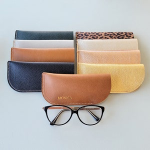May include: A set of ten different colored leather eyeglass cases, with one case featuring the word "MONICA" in gold lettering. The cases are arranged in a row, with the case with the name "MONICA" in the center. A pair of eyeglasses is in front of the cases.