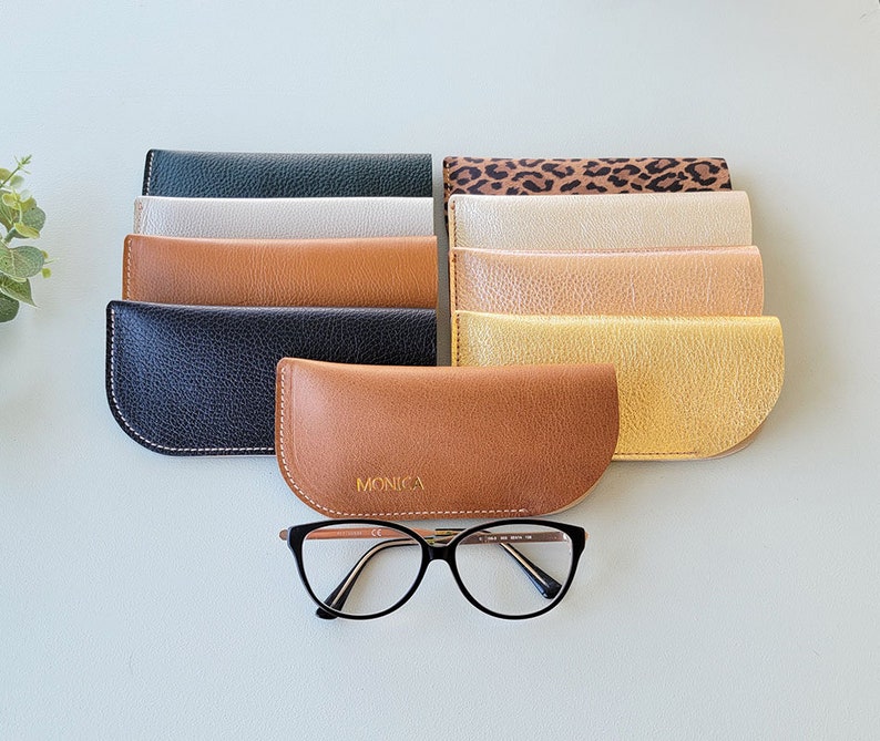 May include: A set of ten different colored leather eyeglass cases, with one case featuring the word "MONICA" in gold lettering. The cases are arranged in a row, with the case with the name "MONICA" in the center. A pair of eyeglasses is in front of the cases.