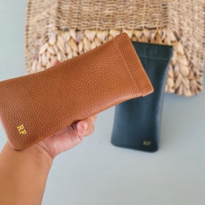 May include: A brown leather glasses case with the initials "RF" in gold lettering. A dark green glasses case is behind it. The cases are rectangular and have a textured surface. The background includes a woven basket.