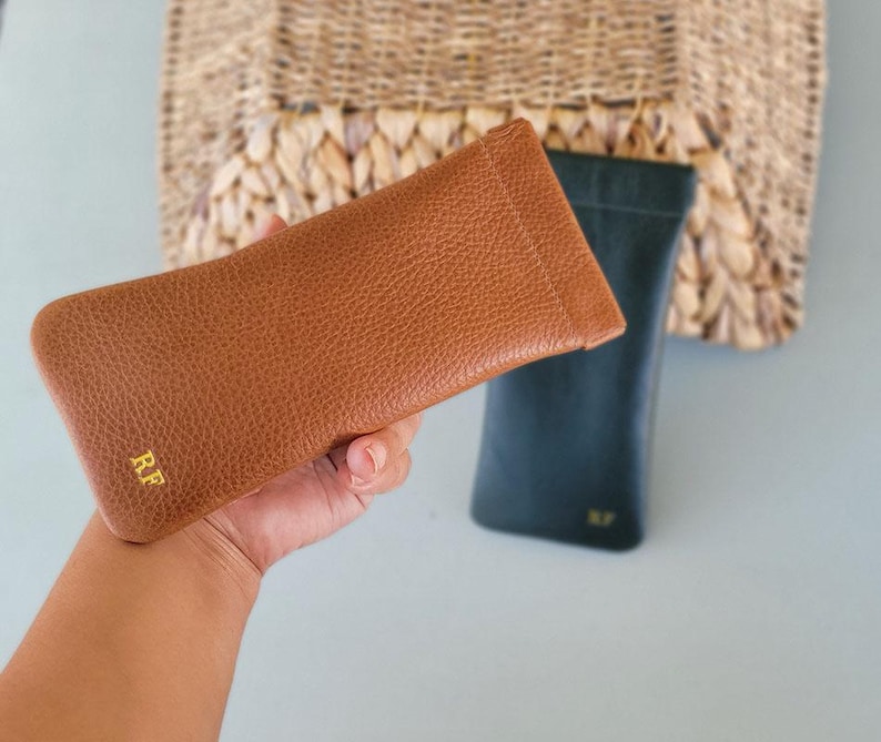 May include: A brown leather glasses case with the initials "RF" in gold lettering. A dark green glasses case is behind it. The cases are rectangular and have a textured surface. The background includes a woven basket.