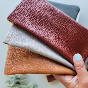 May include: A collection of leather eyeglass cases in various colors, including brown, gray, and burgundy. The cases have a textured surface and a rectangular shape with a gold-colored metal accent at the opening. The cases are held in a hand.