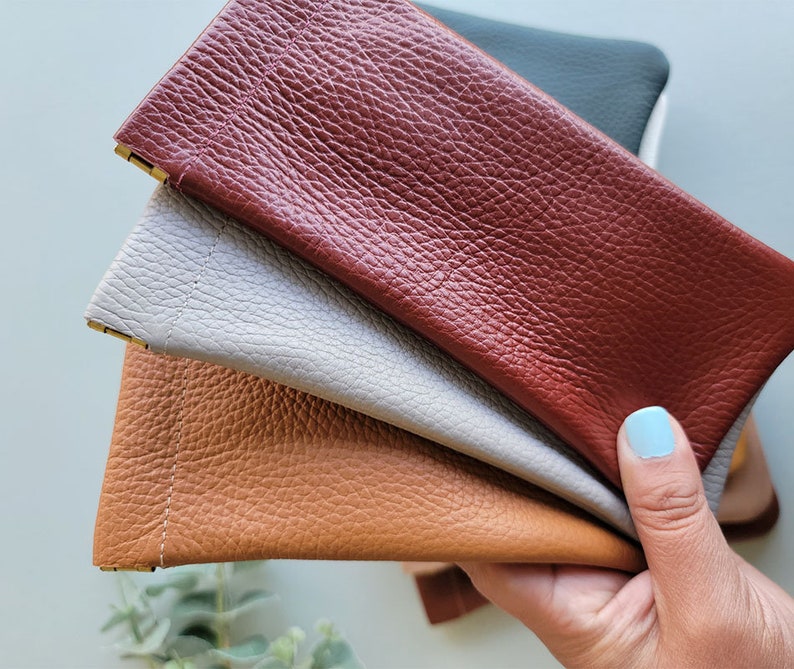 May include: A collection of leather eyeglass cases in various colors, including brown, gray, and burgundy. The cases have a textured surface and a rectangular shape with a gold-colored metal accent at the opening. The cases are held in a hand.