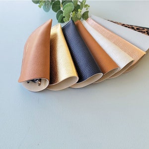 May include: A set of eight leather eyeglass cases in various colors, including brown, gold, black, and white. The cases are lined up in a fan shape, with the brown case at the front.