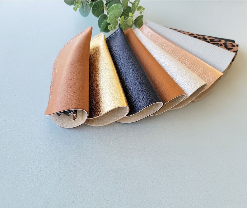 May include: A set of eight leather eyeglass cases in various colors, including brown, gold, black, and white. The cases are lined up in a fan shape, with the brown case at the front.