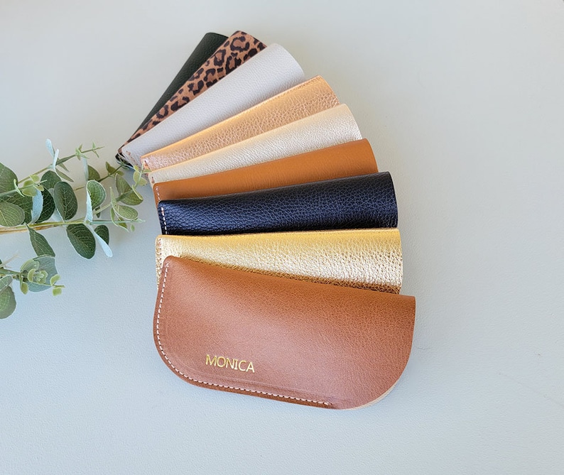 May include: A set of eight different colored leather eyeglass cases. The cases are arranged in a fan shape, with the bottom case being brown and having the name "MONICA" embossed in gold lettering.