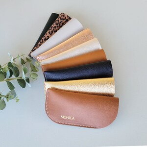 May include: A collection of eight different colored leather eyeglass cases, arranged in a fan shape. The bottom case is brown and has the name "MONICA" embossed in gold lettering.