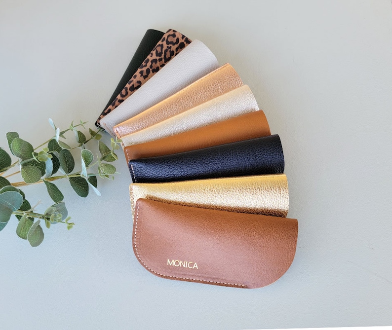 May include: A collection of eight different colored leather eyeglass cases, arranged in a fan shape. The bottom case is brown and has the name "MONICA" embossed in gold lettering.