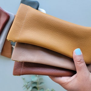 May include: A collection of leather eyeglass cases in various colors, including tan, brown, and gray. The cases are rectangular with a stitched seam and a small metal clasp. The leather has a textured appearance.