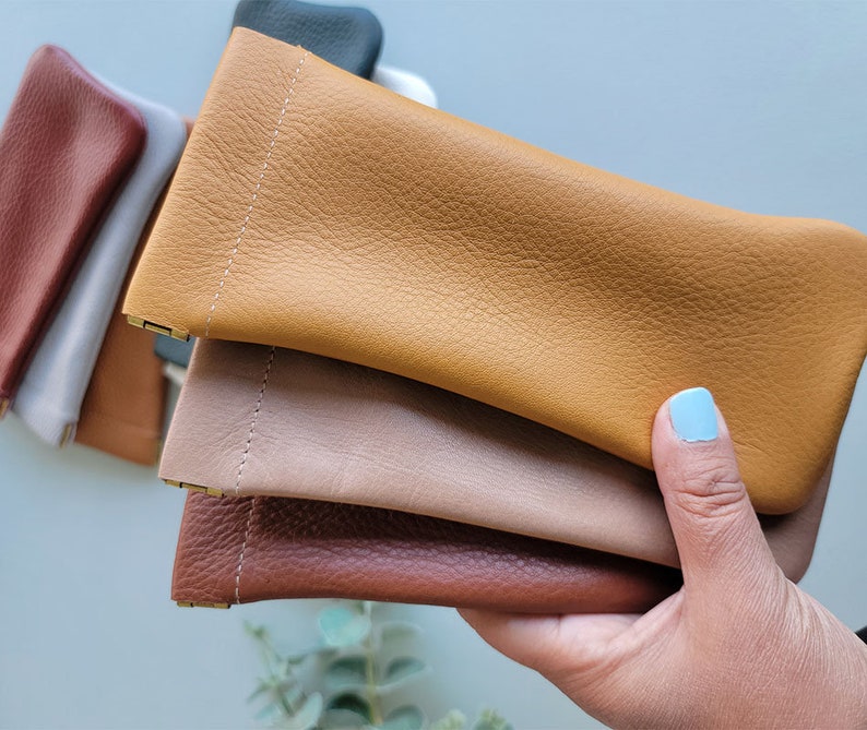 May include: A collection of leather eyeglass cases in various colors, including tan, brown, and gray. The cases are rectangular with a stitched seam and a small metal clasp. The leather has a textured appearance.