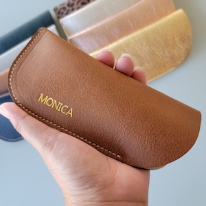 May include: A brown leather glasses case with a gold stitched edge and the word "MONICA" embossed in gold lettering.