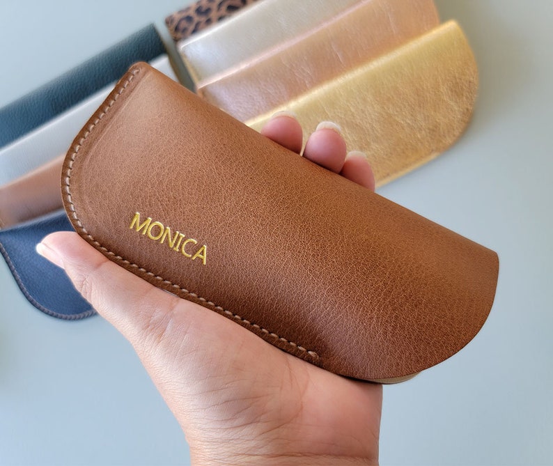 May include: A brown leather glasses case with a gold stitched edge and the word "MONICA" embossed in gold lettering.