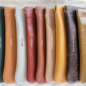 May include: A collection of leather pouches in various colors, including white, green, orange, gray, red, brown, and tan. Each pouch has a stitched top and a brass-colored clasp. The colors are labeled with text.