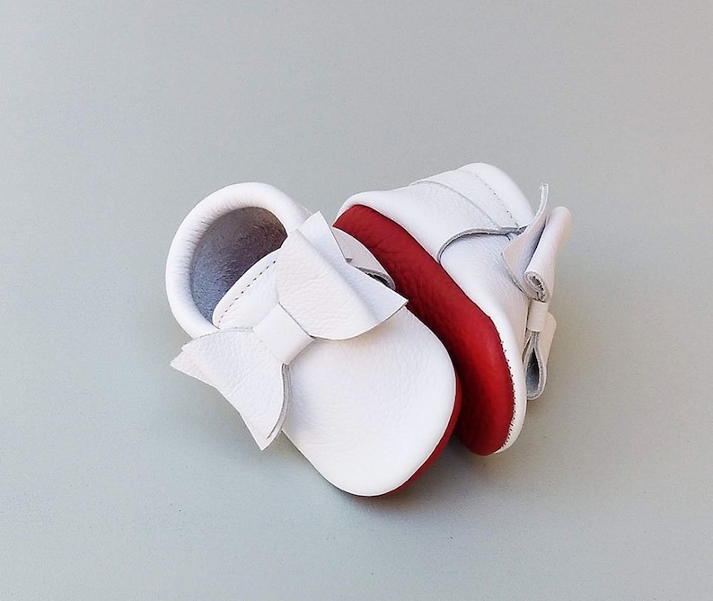 Puede incluir: Un par de zapatos de beb&eacute; de cuero blanco con suelas rojas y un gran lazo en la parte delantera. Los zapatos est&aacute;n dise&ntilde;ados con una suela blanda y una correa para sujetarlos al pie. Est&aacute;n hechos de un material suave y flexible.