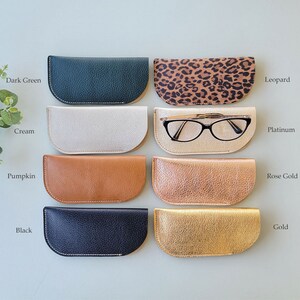 May include: Eight different colored leather eyeglass cases, including dark green, cream, pumpkin, black, leopard print, platinum, rose gold, and gold. The case in the middle row, second from the left, has a pair of eyeglasses inside.