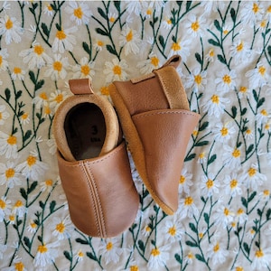 May include: Two pairs of brown leather baby shoes with a suede toe. The shoes are on a white background with a floral pattern.
