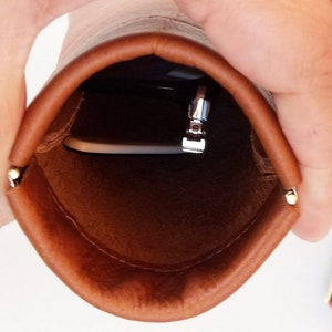 May include: A brown leather pouch with a silver zipper and a small metal accent. The pouch is open, revealing a silver object inside. The pouch is held open by a person's hands.