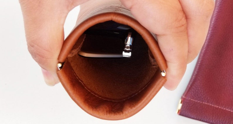 May include: A brown leather pouch with a silver zipper and a small metal accent. The pouch is open, revealing a silver object inside. The pouch is held open by a person's hands.