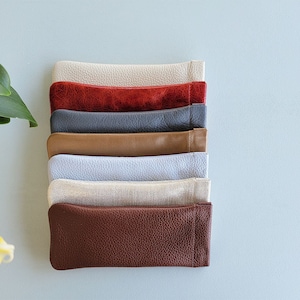 May include: A stack of seven leather eyeglass cases in various colors, including beige, red, gray, brown, light blue, cream, and dark brown.