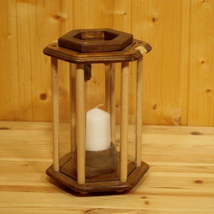 Könnte beinhalten: Eine Kerzenlaterne aus Holz mit einer weißen Kerze im Inneren. Die Laterne hat einen sechseckigen Sockel und einen transparenten Glaseinsatz.