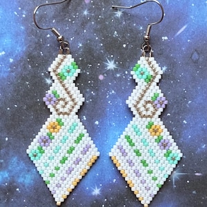 May include: A pair of beaded earrings with a geometric design. The earrings feature white, green, yellow, and purple beads arranged in a diamond shape. The earrings have silver-colored ear wires and a decorative pattern.
