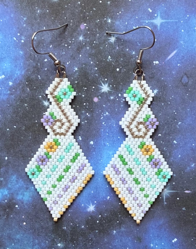 May include: A pair of beaded earrings with a geometric design. The earrings feature white, green, yellow, and purple beads arranged in a diamond shape. The earrings have silver-colored ear wires and a decorative pattern.