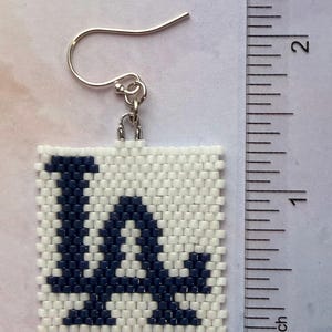 Puede incluir: Pendiente cuadrado de cuentas con las letras "LA" en azul marino sobre fondo blanco. El pendiente, con un gancho plateado, mide aproximadamente 2,5 cm cuadrados.
