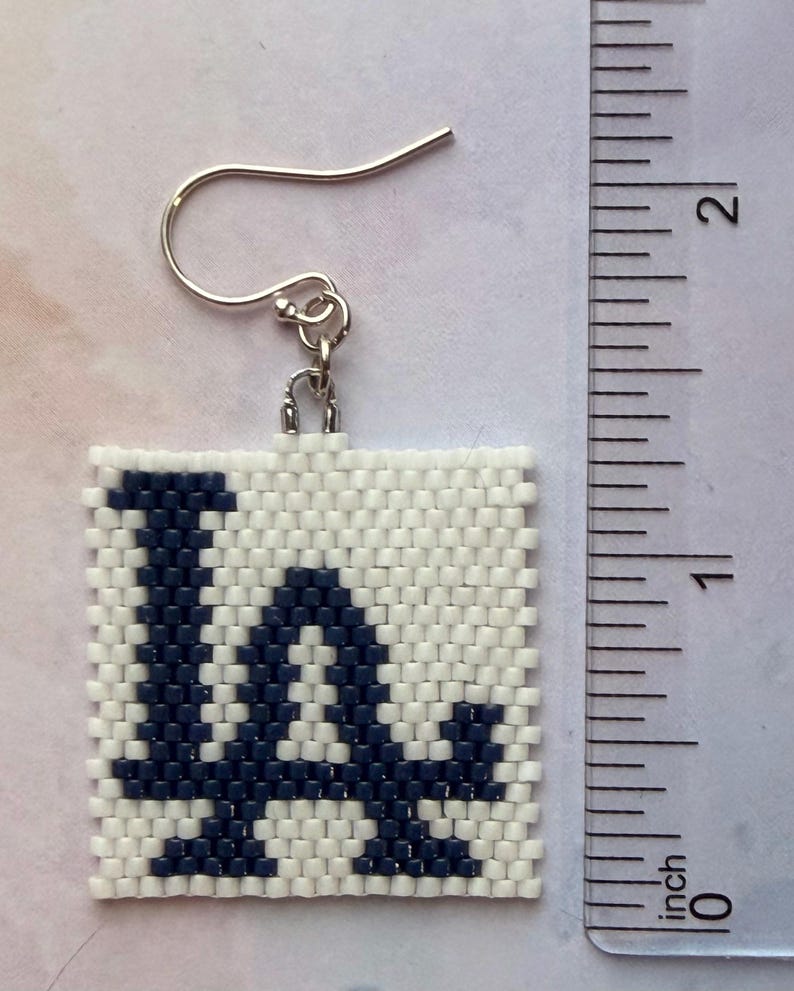 Puede incluir: Pendiente cuadrado de cuentas con las letras "LA" en azul marino sobre fondo blanco. El pendiente, con un gancho plateado, mide aproximadamente 2,5 cm cuadrados.