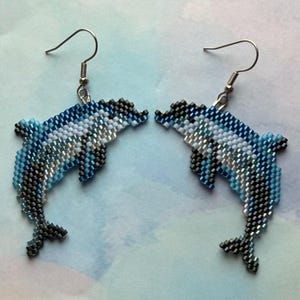 May include: A pair of beaded dolphin earrings. The earrings feature a detailed design with shades of blue, white, and silver beads. The dolphins are suspended from silver-colored fish hook ear wires, creating a nautical theme.
