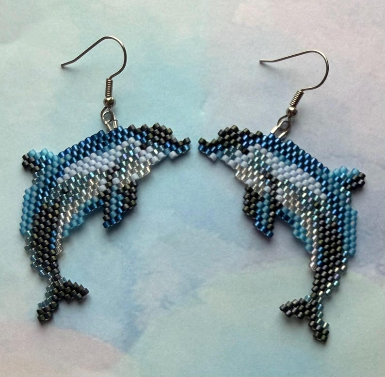 May include: A pair of beaded dolphin earrings. The earrings feature a detailed design with shades of blue, white, and silver beads. The dolphins are suspended from silver-colored fish hook ear wires, creating a nautical theme.