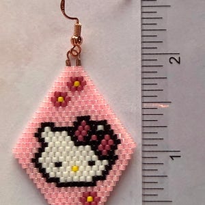 Może przedstawiać: R&oacute;żowy kolczyk z koralik&oacute;w z motywem Hello Kitty. Kolczyk ma kształt rombu z r&oacute;żowym tłem i twarzą Hello Kitty w kolorze białym, czarnym i ż&oacute;łtym. Obecne są r&oacute;wnież małe r&oacute;żowe kwiaty z ż&oacute;łtymi środkami. Kolczyk ma około 2,5 cm długości.