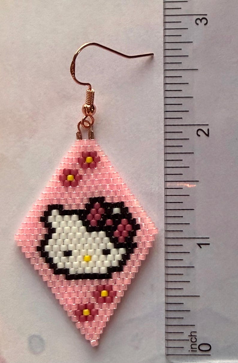 Może przedstawiać: R&oacute;żowy kolczyk z koralik&oacute;w z motywem Hello Kitty. Kolczyk ma kształt rombu z r&oacute;żowym tłem i twarzą Hello Kitty w kolorze białym, czarnym i ż&oacute;łtym. Obecne są r&oacute;wnież małe r&oacute;żowe kwiaty z ż&oacute;łtymi środkami. Kolczyk ma około 2,5 cm długości.