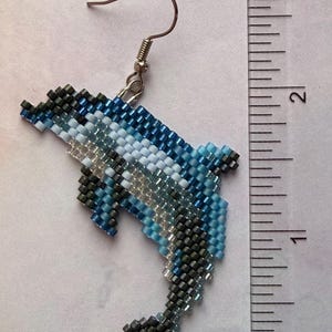 May include: Handmade beaded dolphin earring. The dolphin is crafted with small beads in shades of blue, white, and gray. The earring measures approximately 2 inches in length, including the hook.