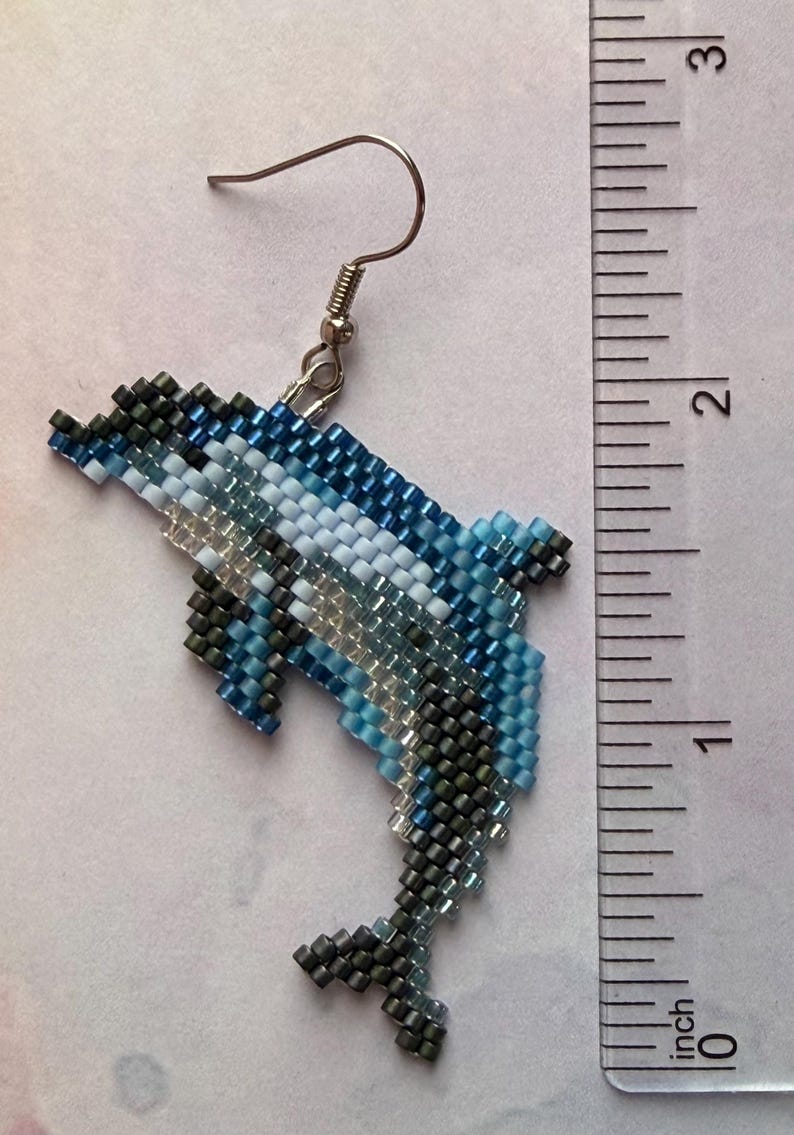 May include: Handmade beaded dolphin earring. The dolphin is crafted with small beads in shades of blue, white, and gray. The earring measures approximately 2 inches in length, including the hook.