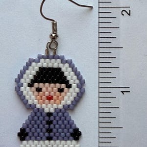 May include: Handmade beaded earring featuring a figure in a hooded parka. The earring is approximately 1.75 inches (4.4 cm) tall. The figure has a black hat, a pink face, and a red mouth. The parka is lavender with black and white accents.