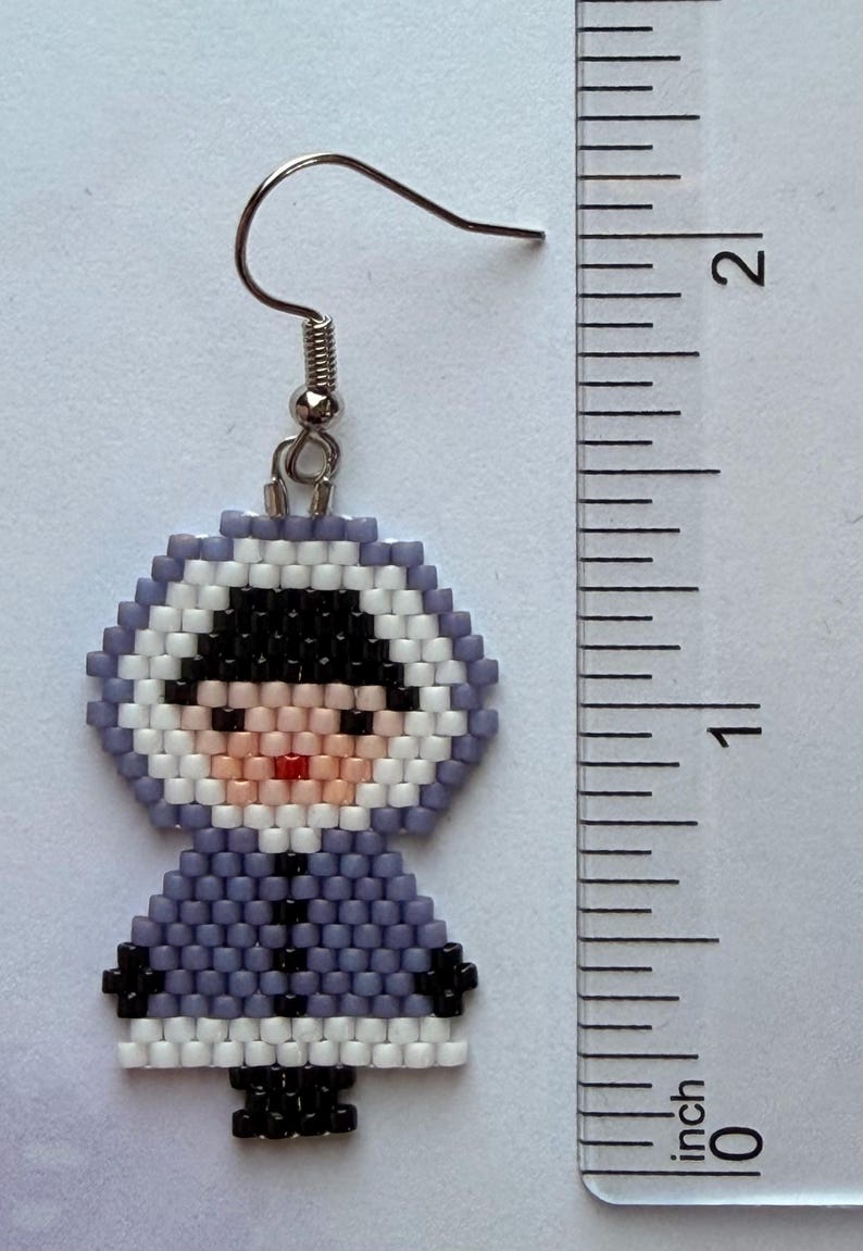 May include: Handmade beaded earring featuring a figure in a hooded parka. The earring is approximately 1.75 inches (4.4 cm) tall. The figure has a black hat, a pink face, and a red mouth. The parka is lavender with black and white accents.