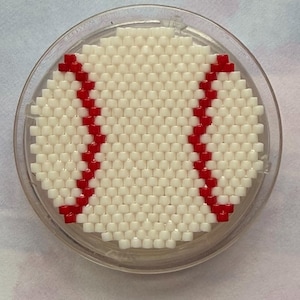 May include: A round coaster featuring a baseball design. The coaster is made of small, off-white beads arranged to resemble a baseball, with red bead stitching. The coaster is encased in a clear, circular frame.