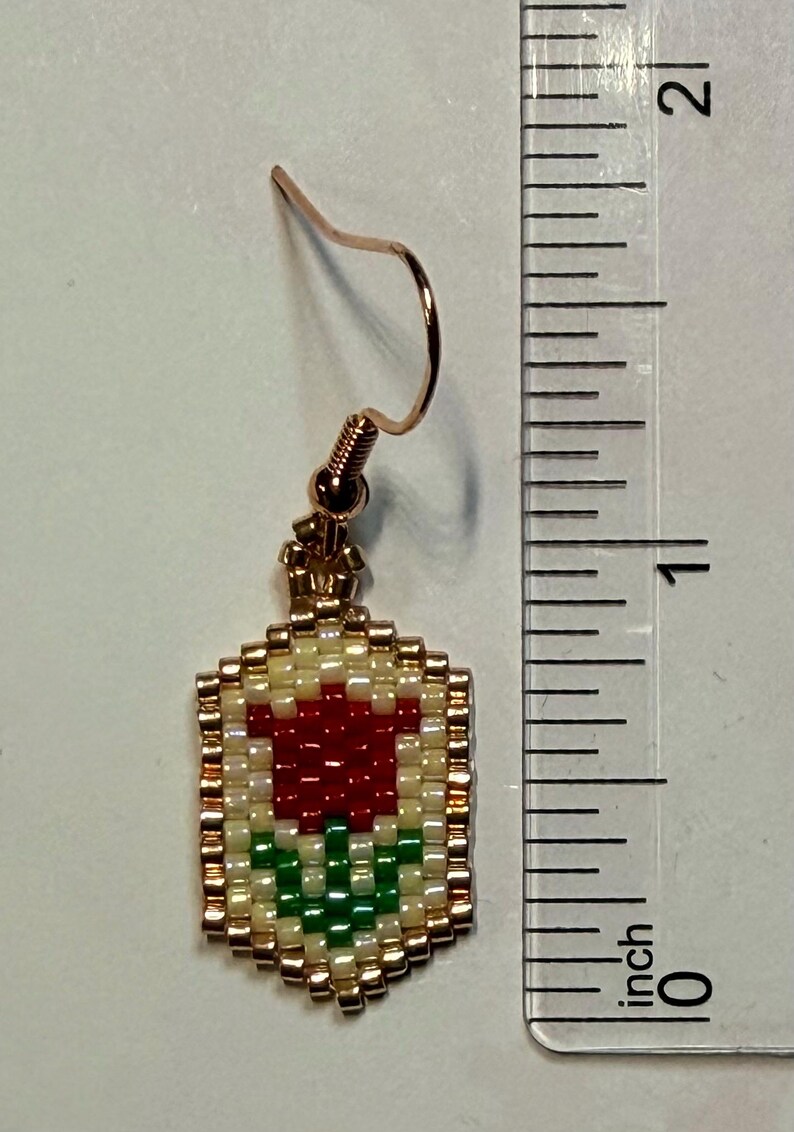 May include: A beaded earring with a rose design. The earring features a red rose with green leaves on a cream background, framed by gold beads. The earring is approximately 1 inch in length.