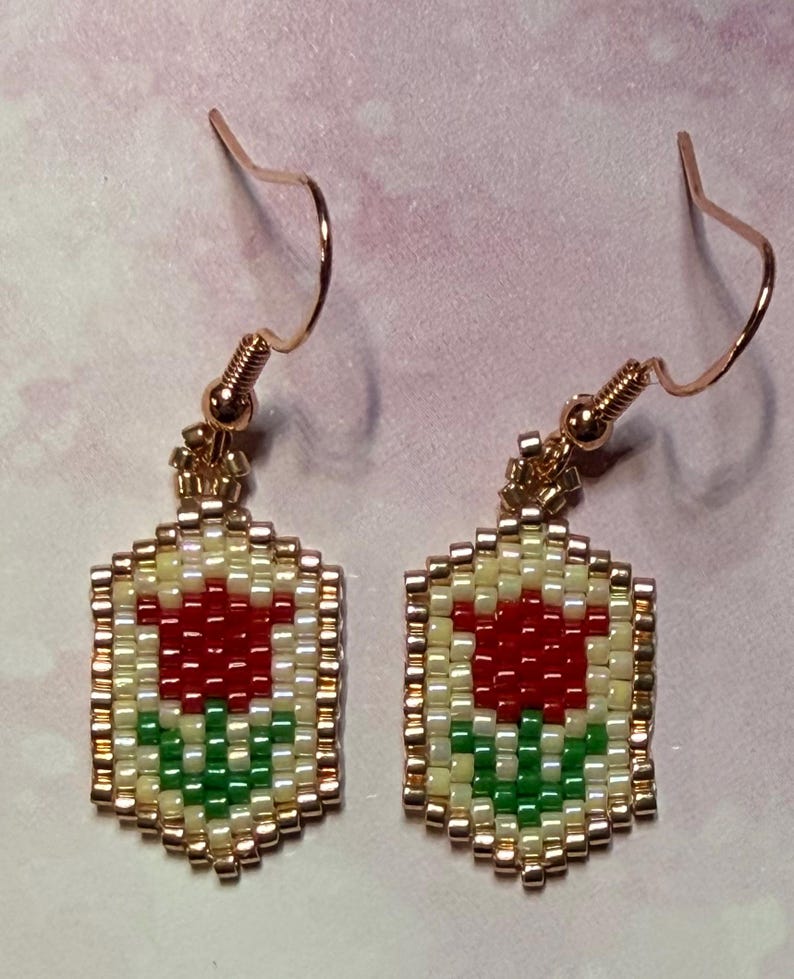 May include: A pair of beaded earrings featuring a red tulip design. The earrings have a gold-colored hook and are made with small, colorful beads. The tulip is set against a cream-colored background, with green leaves and a gold border.