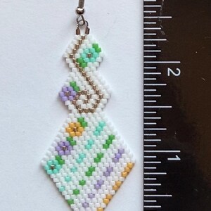 May include: Handmade beaded earrings with a geometric design. The earrings feature a white background with a pattern of green, purple, and yellow beads. The earrings are approximately 2.5 inches long.