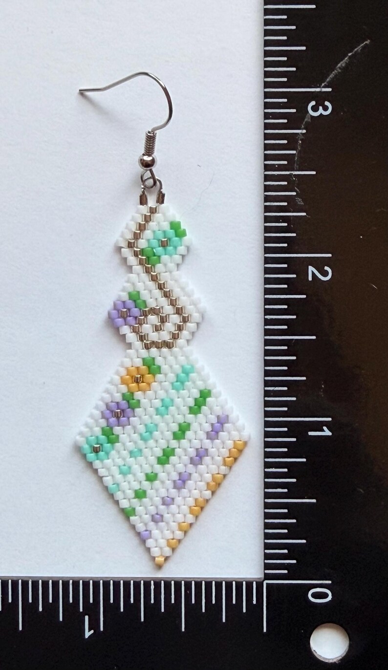 May include: Handmade beaded earrings with a geometric design. The earrings feature a white background with a pattern of green, purple, and yellow beads. The earrings are approximately 2.5 inches long.