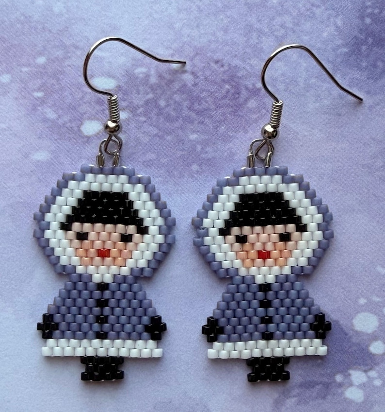 May include: A pair of handmade beaded earrings featuring a figure in a hooded parka. The earrings are primarily lavender, black, white, and peach. The figure has a black hat, a peach face, and a red mouth. The earrings hang from silver-colored fish hook ear wires.