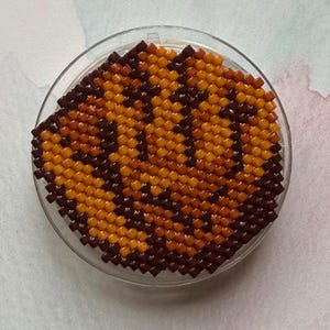 May include: A round, beaded coaster with a brown and orange pattern, set inside a clear, circular holder. The coaster's design features a repeating pattern of squares, creating a textured appearance. The color scheme is warm, with shades of brown and orange.