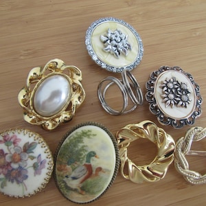 May include: A collection of seven decorative items including circular and oval brooches or scarf slides. Features include a faux pearl in a gold frame, a floral painted disc, a mallard duck illustration, and metallic twisted or filigree designs.