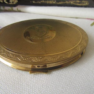 May include: A gold-coloured, oval compact with a textured surface and decorative border. The compact features a shield emblem in the centre. The clasp is visible on the bottom edge. The compact is on a white surface.