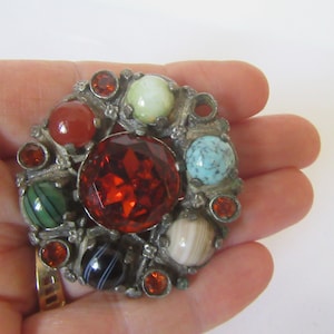 May include: A silver brooch featuring a red, faceted gemstone at its centre, surrounded by smaller, round gemstones in a variety of colours, including green, blue, and brown.