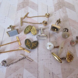 May include: A collection of 12 gold and silver cufflinks and tie tacks. The cufflinks feature a variety of designs, including a square with a geometric pattern, a pair of oval cufflinks with a winged angel design, and a pair of oval cufflinks with a pearl-like center. The tie tacks feature a variety of designs, including a gold chain with a small ball at the end, a gold chain with a small bar at the end, and a silver chain with a small bar at the end.