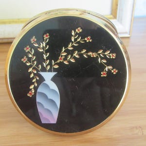 May include: Round black compact with a gold rim. The top features a gold and red floral design with a white and gray vase. The compact is likely used for cosmetics and has a vintage aesthetic.