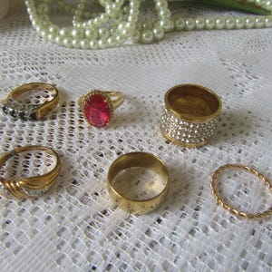 Puede incluir: Una colección de anillos de oro con varios diseños, incluyendo una banda con piedras transparentes, un anillo con una gran piedra roja y una banda gruesa cubierta de pequeñas piedras transparentes. También hay un anillo de oro trenzado y una banda de oro lisa.