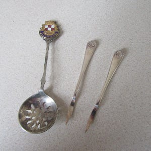 May include: Three silver spoons, one with a decorative handle and a perforated bowl, and two with ornate handles. The spoon with the decorative handle has a small enamel crest with the words "City of Westminster" on it.